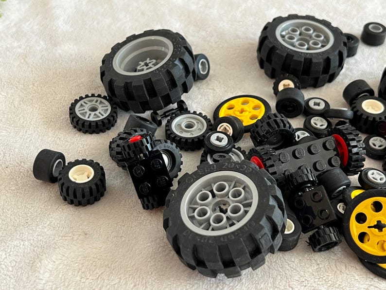 Op de afbeelding: Een verzameling zwarte speelgoedbanden in verschillende maten, sommige met grijze of gele velgen. Er zijn ook verschillende zwarte Lego-stenen aanwezig, sommige met rode accenten. De banden liggen verspreid op een wit oppervlak.