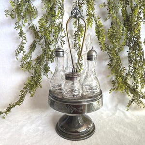 Könnte beinhalten: Ein Vintage-Gewürzset aus versilbertem Metall mit vier durchsichtigen Glasflaschen und einem zentralen Shaker. Das Set enthält einen dekorativen Griff und steht auf einem Sockel. Grünzeug hängt im Hintergrund.