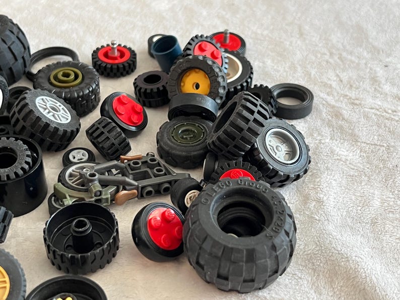 May include: A collection of various black toy tires, wheels, and related components. Some wheels have red or yellow centers. A small toy motorcycle is also present. The tires vary in size and tread pattern.