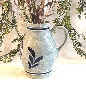 May include: A gray ceramic pitcher with a handle, decorated with a dark blue leaf design and a dark blue band around the top and bottom. The pitcher is filled with dried grasses and stems. Decorative greenery is in the background.