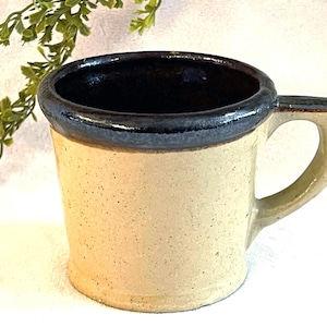 May include: A handmade ceramic mug with a speckled beige exterior and a dark brown interior and rim. The mug has a rounded handle and a slightly flared shape. The mug is a neutral color and would be suitable for coffee or tea.