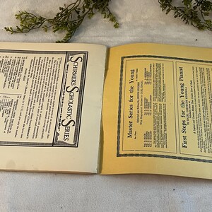 May include: Open vintage book pages with text, featuring "SCHIRMER'S SCHOLASTIC SERIES" and "Master Series for the Young." The pages are cream-colored with black text and borders. Greenery is visible in the background.