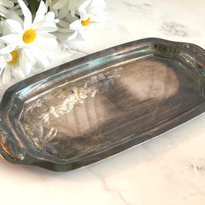 May include: A rectangular silver-toned metal serving tray with rounded corners and integrated handles on each end. The surface of the tray features a reflective, polished finish with visible signs of tarnish and a subtle floral pattern etched into the metal.
