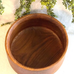 May include: A round, wooden bowl with a rich, brown color. The bowl has a smooth interior and a slightly textured exterior. Greenery is visible in the background. This bowl is suitable for serving salads or displaying decorative items.