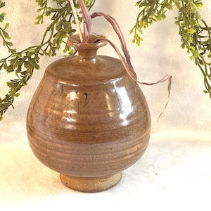 May include: A handcrafted, brown ceramic vase with a rounded body and a narrow neck. The vase has a textured surface with horizontal lines and a small, circular base. A few dried stems are placed inside the vase. Decorative greenery is in the background.