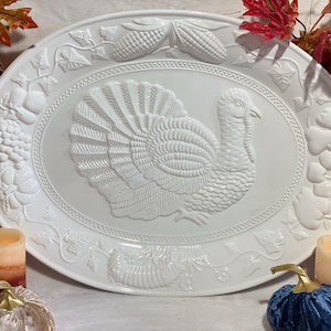 Puede incluir: Plato ovalado blanco para servir con un diseño en relieve de un pavo, maíz, uvas y otros elementos de la cosecha. El plato está rodeado de decoraciones con temática otoñal, incluyendo velas y calabazas decorativas.