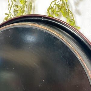 Może przedstawiać: Zbliżenie czarnej tacy do serwowania z bordowym obramowaniem i zużytą, postarzaną krawędzią. Taca ma lekko zakrzywiony kształt i gładką powierzchnię. Tło zawiera zieleń.