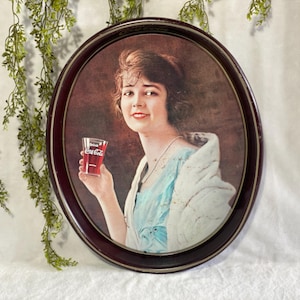 Może przedstawiać: Vintage owalna reklama Coca-Coli przedstawiająca kobietę trzymającą szklankę Coca-Coli. Obraz jest oprawiony w ciemnobrązową owalną ramę. Kobieta ma na sobie niebieską sukienkę i białą szal. Szklanka ma logo Coca-Coli.