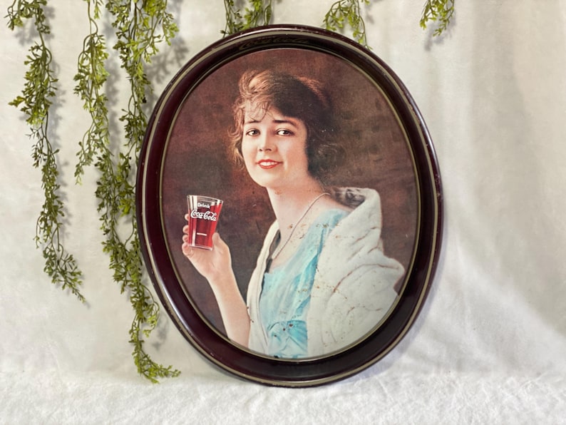 Może przedstawiać: Vintage owalna reklama Coca-Coli przedstawiająca kobietę trzymającą szklankę Coca-Coli. Obraz jest oprawiony w ciemnobrązową owalną ramę. Kobieta ma na sobie niebieską sukienkę i białą szal. Szklanka ma logo Coca-Coli.