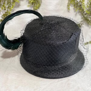 May include: A black hat with a rounded crown and a wide brim, adorned with a dark green feather and a fine black netting veil. The hat has a textured, woven appearance, suggesting a straw or similar material.