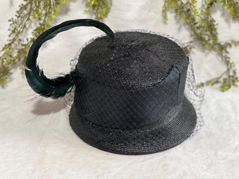 May include: A black hat with a rounded crown and a wide brim, adorned with a dark green feather and a fine black netting veil. The hat has a textured, woven appearance, suggesting a straw or similar material.