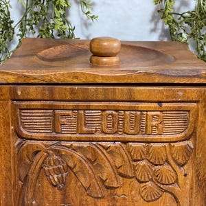 May include: A wooden flour container with a lid and a small knob. The word "FLOUR" is carved into the front of the container. The container has carved floral and leaf designs. The wood has a warm, brown tone.
