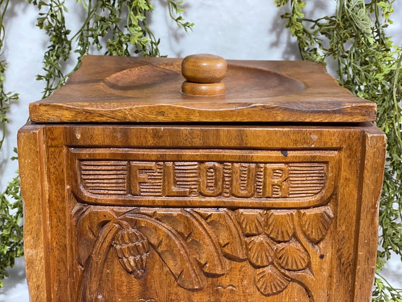 May include: A wooden flour container with a lid and a small knob. The word "FLOUR" is carved into the front of the container. The container has carved floral and leaf designs. The wood has a warm, brown tone.