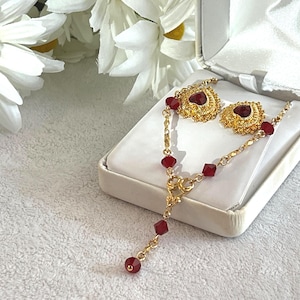 Op de afbeelding: Een goudkleurige sieradenset met een ketting en oorbellen met rode edelsteenaccenten. De oorbellen en ketting worden gepresenteerd in een witte sieradendoos. De set is geaccentueerd met rode, vierkant geslepen edelstenen.