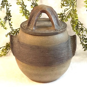 May include: A handcrafted ceramic jar with a lid. The jar is a muted tan color with a darker brown band around the middle. The lid has a handle and a textured surface. The jar has two small handles on the sides.