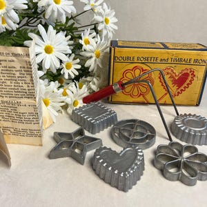 May include: A vintage Double Rosette and Timbale Iron set featuring a yellow box with red heart graphics, a red-handled metal tool, and five assorted aluminum molds in star, heart, round, and flower shapes, accompanied by a printed instruction sheet.