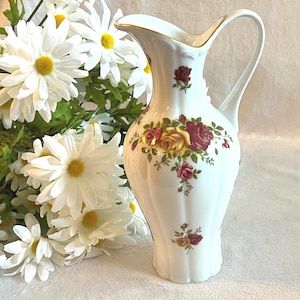 Peut inclure: Pichet en porcelaine blanche avec un bord doré et une anse. Le pichet est orné d'un motif floral de roses rouges et jaunes. Un bouquet de marguerites blanches à cœur jaune est à côté du pichet.