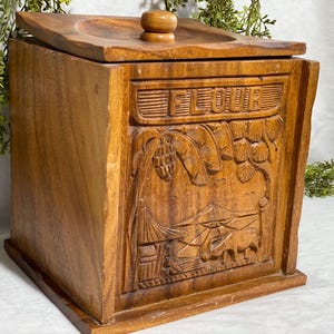 May include: Wooden flour container with a lid and a carved design. The word "FLOUR" is carved across the top. The front of the container features a detailed relief carving of a landscape with buildings and animals.