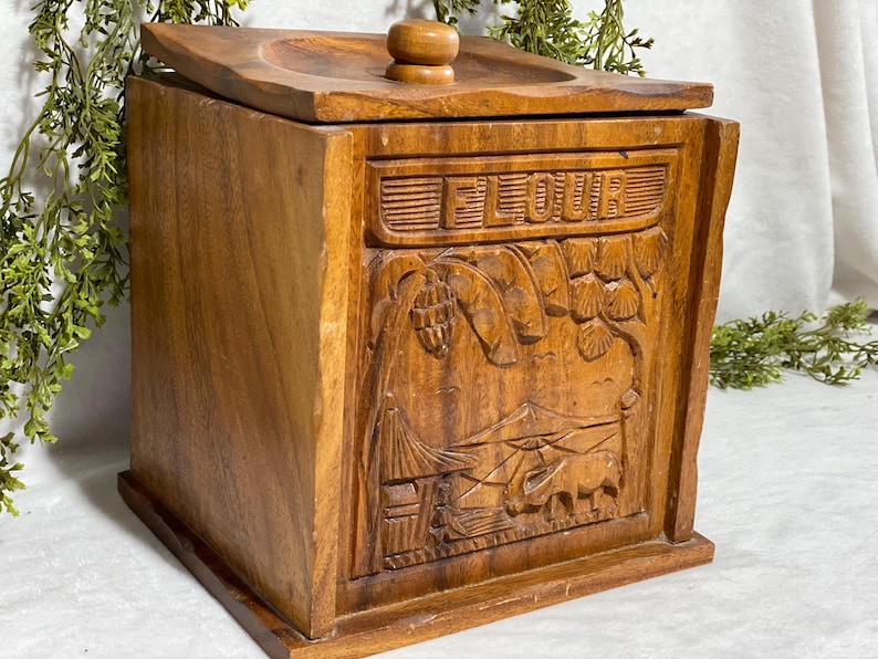 May include: Wooden flour container with a lid and a carved design. The word "FLOUR" is carved across the top. The front of the container features a detailed relief carving of a landscape with buildings and animals.