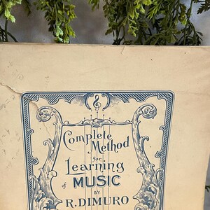 May include: An antique book cover with the title "Complete Method for Learning Music" in blue script. The cover has an ornate blue border and is set against a beige background. The book is slightly worn.
