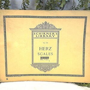May include: A vintage book cover with the text "Schirmer's Library" and "Herz Scales." The cover is a light yellow color with a black decorative border and text. The book is slightly worn, suggesting it is an antique.