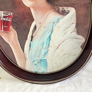 Może przedstawiać: Vintage reklama Coca-Coli w ciemnej owalnej ramie. Obraz przedstawia osobę trzymającą szklankę Coca-Coli. Osoba ma na sobie niebieski top i białą szal. Rama ma ciemnobrązowe wykończenie.
