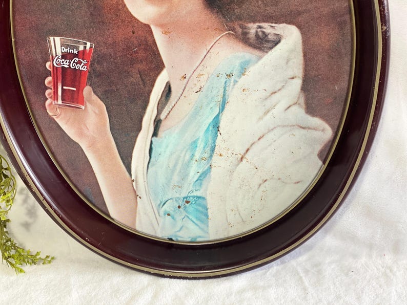 Może przedstawiać: Vintage reklama Coca-Coli w ciemnej owalnej ramie. Obraz przedstawia osobę trzymającą szklankę Coca-Coli. Osoba ma na sobie niebieski top i białą szal. Rama ma ciemnobrązowe wykończenie.