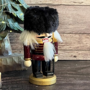 Könnte beinhalten: Eine hölzerne Nussknacker-Figur mit einer hohen schwarzen Kunstfellmütze, einer dunkelroten Jacke mit Goldverzierungen und einer schwarzen Hose. Die Figur hat einen Bart und Haare aus weißem Kunstfell, bemalte Gesichtszüge und steht auf einem runden goldenen Sockel.