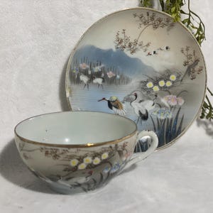 May include: A vintage teacup and saucer set. The cup and saucer are decorated with a serene landscape featuring cranes, flowers, and a mountain range. The set has a gold trim, adding a touch of elegance to the design. The cup has a handle.