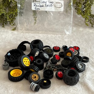 May include: A collection of black Lego tires of various sizes, some with yellow, red, and white accents. The tires are scattered on a white surface, with a plastic bag in the background that reads "Lego Tires Random Lot #3 8oz."