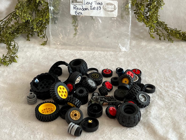 May include: A collection of black Lego tires of various sizes, some with yellow, red, and white accents. The tires are scattered on a white surface, with a plastic bag in the background that reads "Lego Tires Random Lot #3 8oz."
