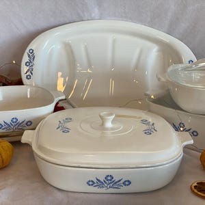 Może przedstawiać: Zestaw białych ceramicznych naczyń do pieczenia z niebieskimi wzorami kwiatowymi. Zestaw zawiera dużą owalną formę, mniejszą owalną formę, okrągłą formę z pokrywką i prostokątną formę.