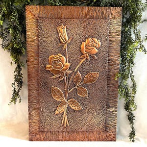 May include: A rectangular copper wall plaque featuring a raised relief design of three roses on a single stem. The central panel has a textured finish, surrounded by a wide, deeply etched border with a dark, metallic bronze-toned patina.