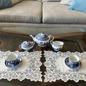 May include: A white porcelain tea set featuring a blue willow pattern, including a teapot, a lidded sugar bowl, a creamer, and two teacups with matching saucers, all arranged on two rectangular white lace placemats with floral borders.