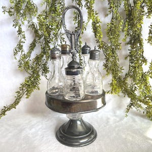 May include: A vintage silver-plated condiment set with six clear glass bottles and jars. The set includes a central jar and five smaller bottles, each with a silver lid. The set is on a tiered stand with a handle, and is surrounded by green foliage.