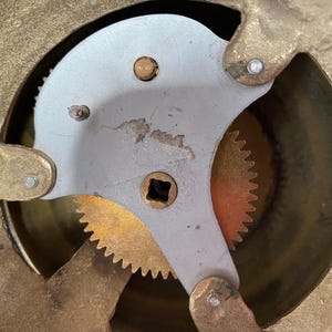 Op de afbeelding: Close-up van een messing uurwerk met een zilveren tandwiel en een messing tandwiel met tanden. De tandwielen zijn zichtbaar door een ronde opening in de wijzerplaat van de klok.