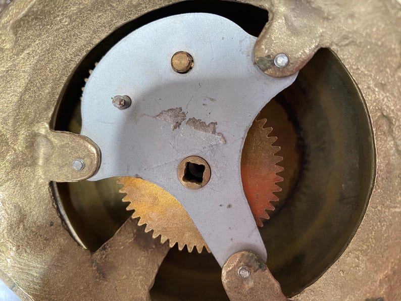 Op de afbeelding: Close-up van een messing uurwerk met een zilveren tandwiel en een messing tandwiel met tanden. De tandwielen zijn zichtbaar door een ronde opening in de wijzerplaat van de klok.