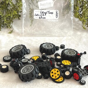 Op de afbeelding: Een verzameling zwarte speelgoedbanden in verschillende maten, sommige met gele en rode accenten, verspreid over een witte ondergrond. Een doorzichtige plastic zak met de tekst "Leap Tires" en "Sets of 4" is zichtbaar.