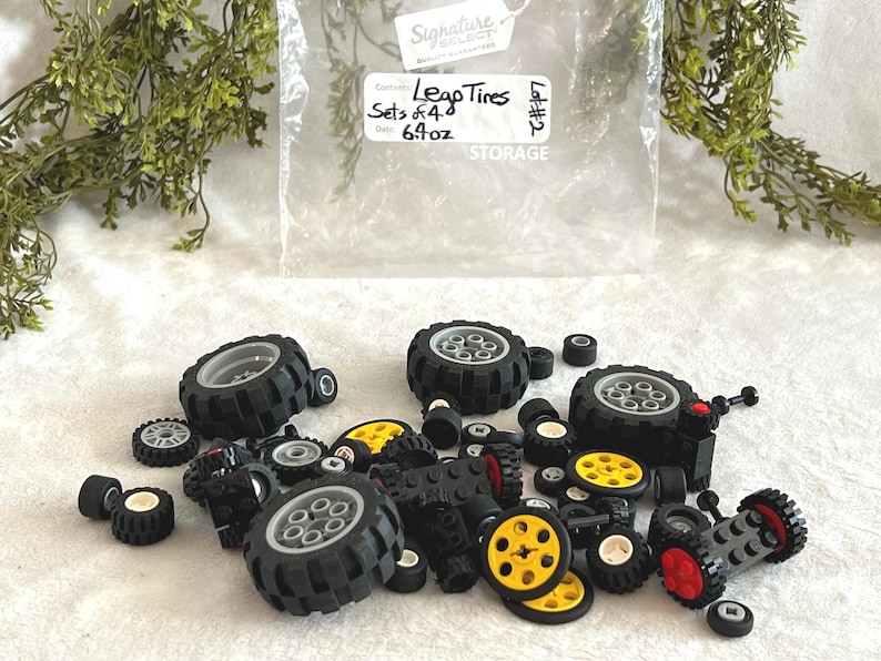 Op de afbeelding: Een verzameling zwarte speelgoedbanden in verschillende maten, sommige met gele en rode accenten, verspreid over een witte ondergrond. Een doorzichtige plastic zak met de tekst "Leap Tires" en "Sets of 4" is zichtbaar.