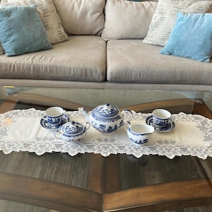 May include: A white ceramic tea set featuring a blue willow pattern, including a teapot, a lidded sugar bowl, a creamer, and two teacups with matching saucers, all arranged on a white lace table runner with intricate floral edges.