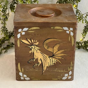 Puede incluir: Un azucarero de madera vintage con tapa. El azucarero es marrón con un diseño de gallo pintado a mano en dorado y detalles blancos. La palabra "SUGAR" es visible en la parte delantera. El azucarero es de forma cuadrada.