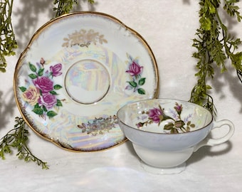 Puede incluir: Un juego de taza y platillo de té vintage. La porcelana blanca presenta detalles iridiscentes y está adornada con rosas rosas y amarillas. Los bordes están ribeteados en dorado. La taza tiene un asa y se apoya en una base blanca.