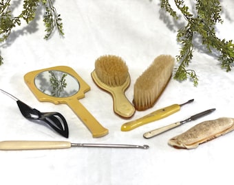 Può includere: Una collezione di strumenti per la cura della persona vintage, tra cui uno specchio a mano giallo, spazzole e strumenti per manicure. Il set presenta una varietà di forme e dimensioni, con manici e setole di colore chiaro. Gli articoli sono disposti su una superficie bianca.
