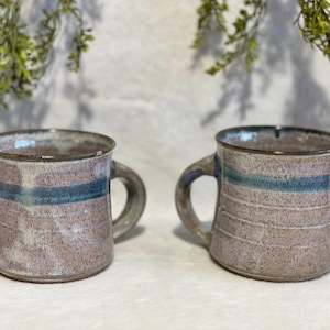 May include: Two handmade ceramic mugs. Each mug has a tan base color with a horizontal blue stripe and a brown rim. The mugs have a curved handle and are displayed on a white surface.