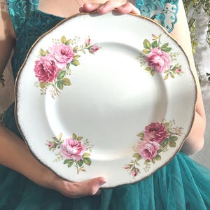 May include: White ceramic plate with a scalloped edge and gold trim. The plate is decorated with pink roses and green leaves. A classic, vintage-style plate.