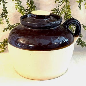 May include: A two-tone ceramic crock with a dark brown top and a white base. The crock has a matching lid and a curved handle. The design is reminiscent of vintage kitchenware.