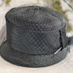 May include: A black hat with a rounded crown and a small brim. The hat is adorned with a black veil and a decorative band. A silver-colored metal bar is attached to the band. The hat is made of woven material.
