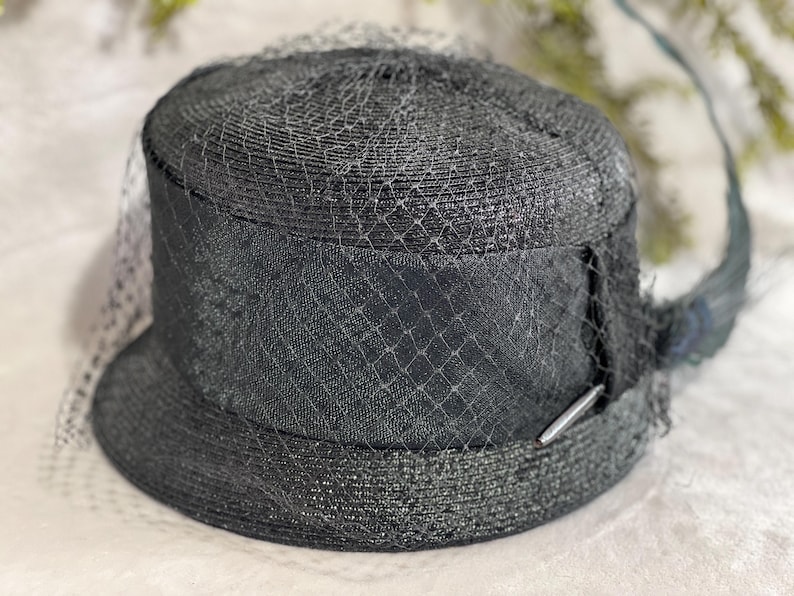 May include: A black hat with a rounded crown and a small brim. The hat is adorned with a black veil and a decorative band. A silver-colored metal bar is attached to the band. The hat is made of woven material.