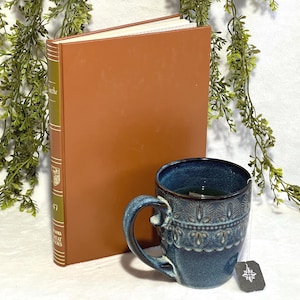 Może przedstawiać: Niebieski ceramiczny kubek z dekoracyjnym wzorem i torebką herbaty, obok brązowej książki. Książka ma złoty grzbiet z tekstem. Zieleń jest ułożona wokół przedmiotów, tworząc przytulną, zachęcającą scenę.