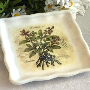 Może przedstawiać: Kwadratowy, kremowy ceramiczny talerz z falistą krawędzią. Na środku znajduje się botaniczna ilustracja szałwii z niebieską wstążką, napisem "Salvia" i skryptem. Stokrotki są w tle.